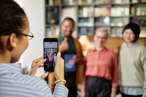 A person is holding up a smartphone to take a photo or video of three smiling people posing together indoors. The group stands in front of bookshelves, with one person giving a thumbs-up.