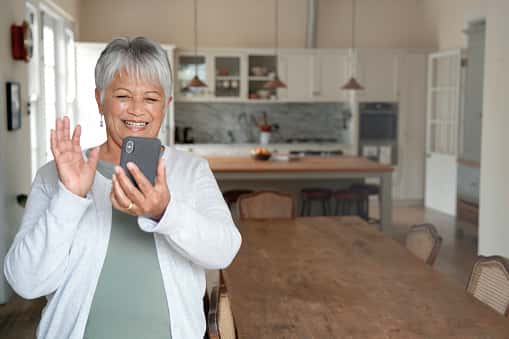 A smiling older woman with short gray hair waves at her smartphone during a video call. She is standing in a bright kitchen with a large wooden dining table in the foreground.