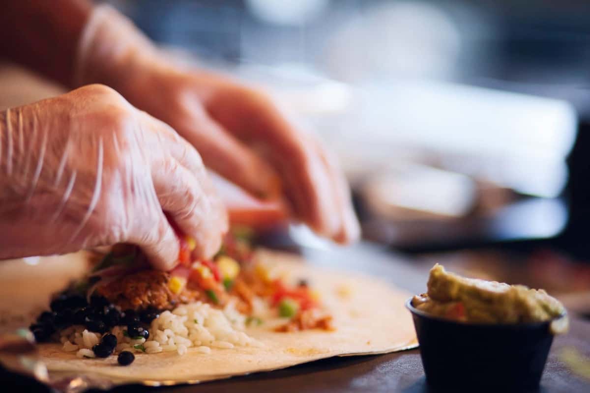 Hands wearing food-safe gloves assembling a fresh Burrito Famous burrito with seasoned meat, rice, black beans, salsa, and guacamole on a tortilla in the restaurant kitchen.