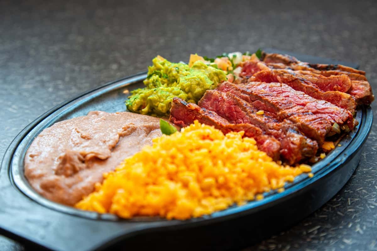 Mexican carne asada plate featuring grilled steak slices, vibrant yellow Mexican rice, creamy refried beans, fresh guacamole, and pico de gallo, all served in a black oval takeout container on a dark textured surface.