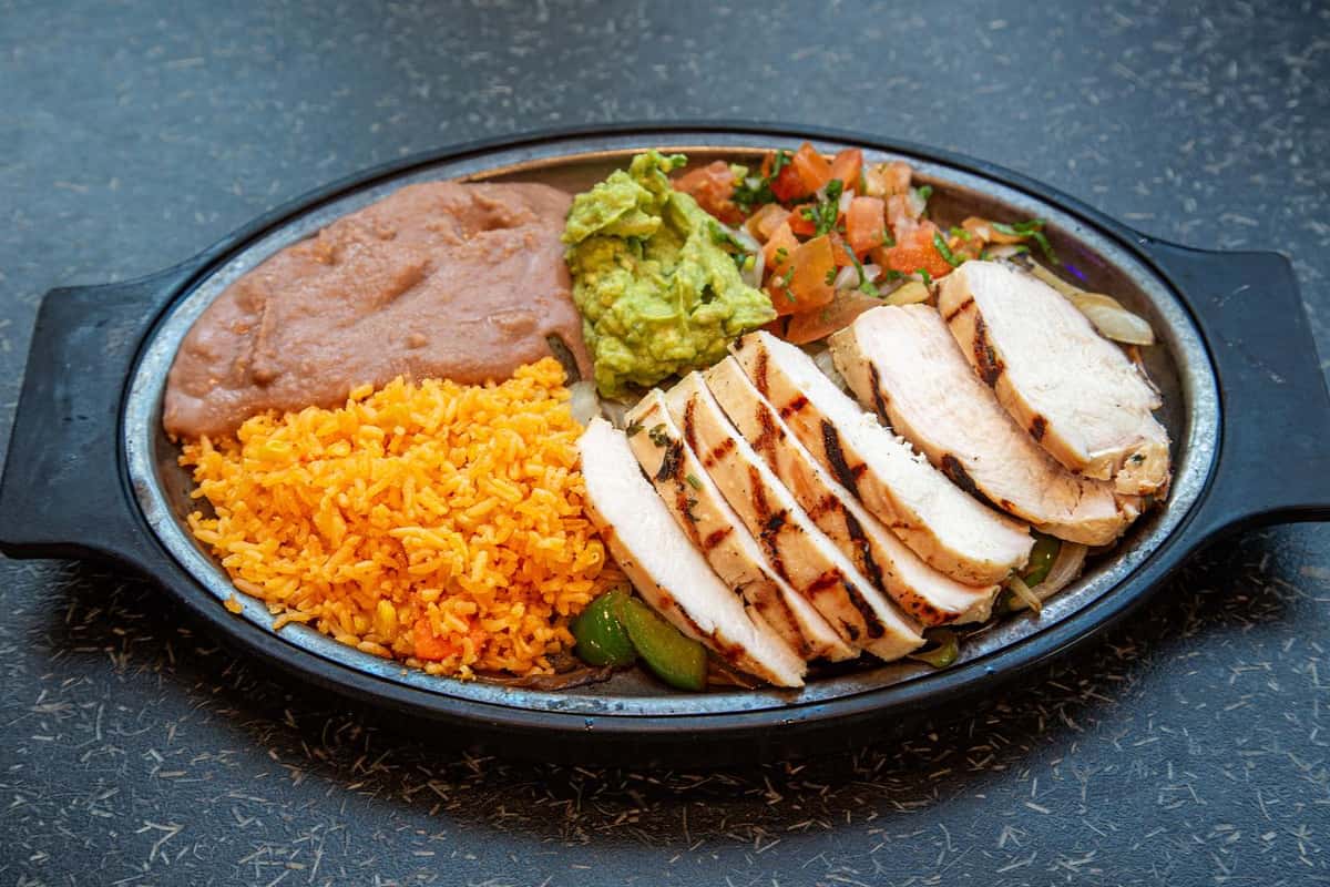 Grilled chicken fajita plate with sliced chicken breast served alongside Mexican rice, refried beans, fresh guacamole, and pico de gallo on a sizzling black oval platter.
