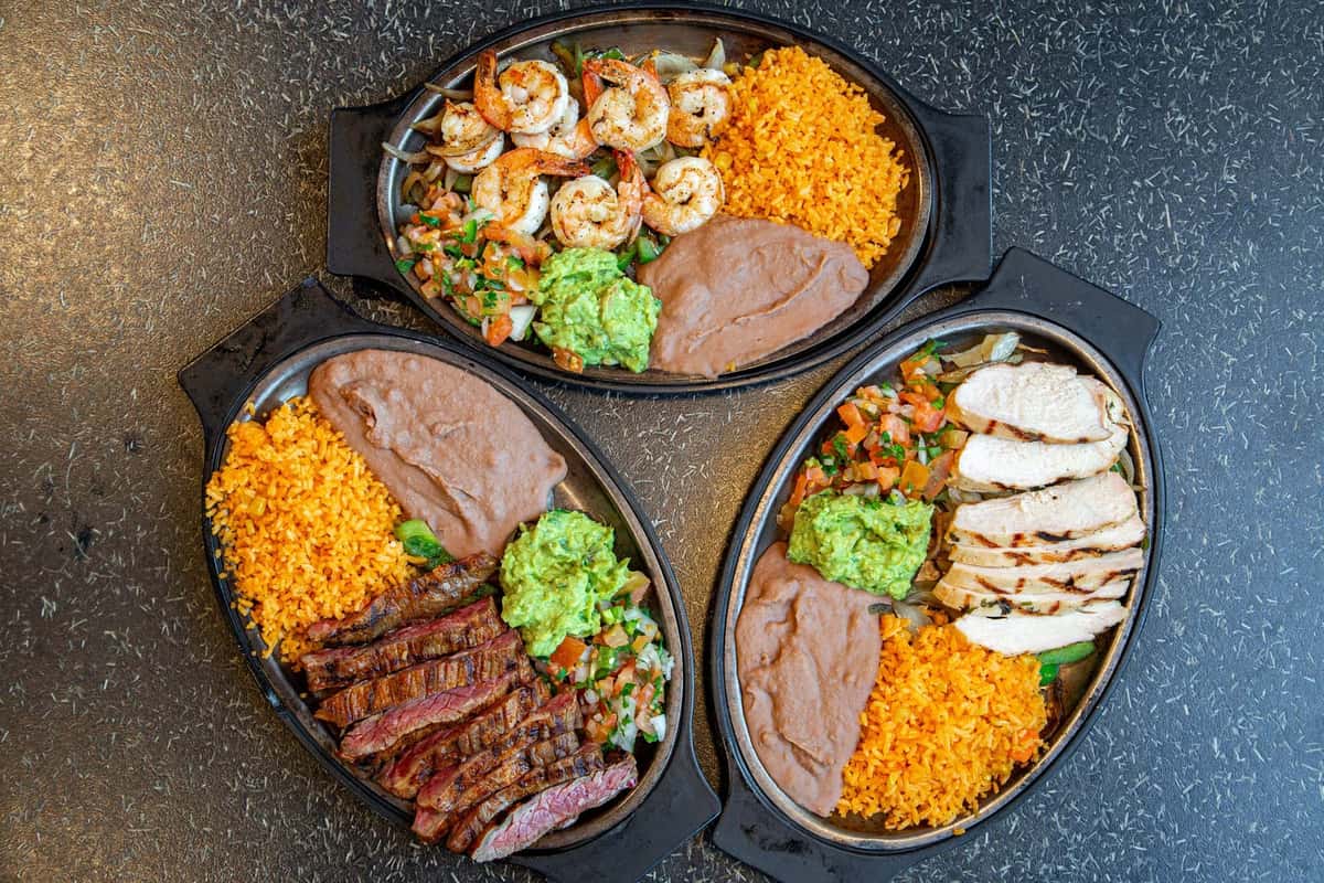 Trio of sizzling Mexican combo plates featuring grilled shrimp, carne asada, and chicken breast, each served with refried beans, Mexican rice, fresh guacamole, and pico de gallo in black oval containers on a speckled dark surface.