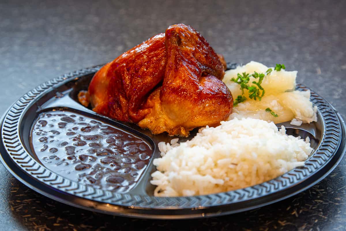 Juicy rotisserie chicken served with white rice, black beans, and yuca topped with parsley in a black takeout plate.