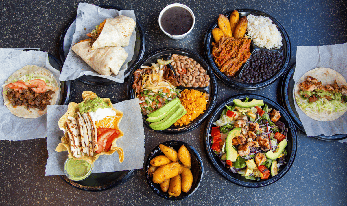 Colorful spread of Mexican food including carne asada tacos, chicken burritos, tostada salad with grilled chicken, rice and beans plates, fried plantains, and a fresh grilled chicken salad with avocado, served on black plates over a dark table surface.