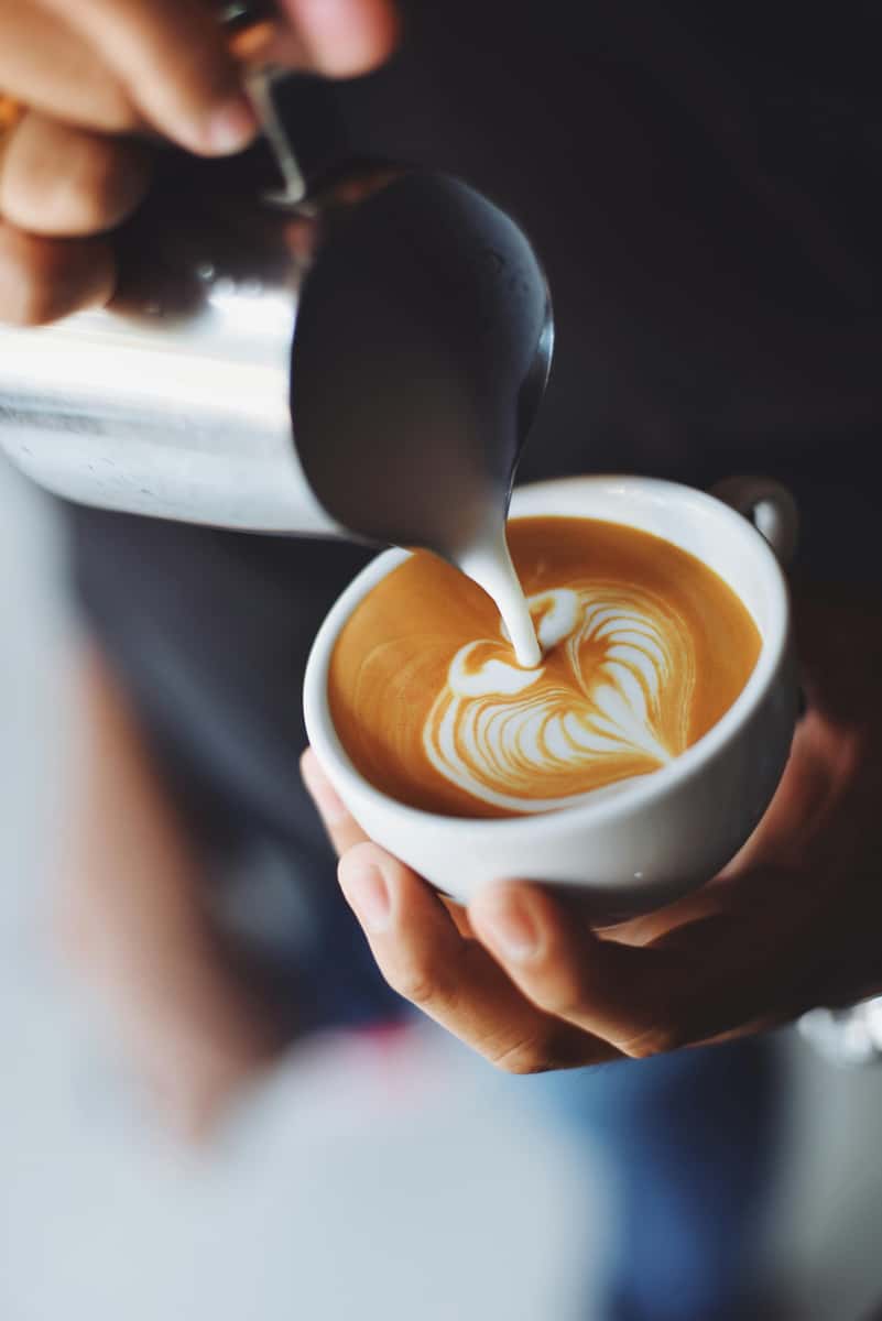 Milk being poured into a cup of coffee creating heart-shaped latte art, held in both hands.