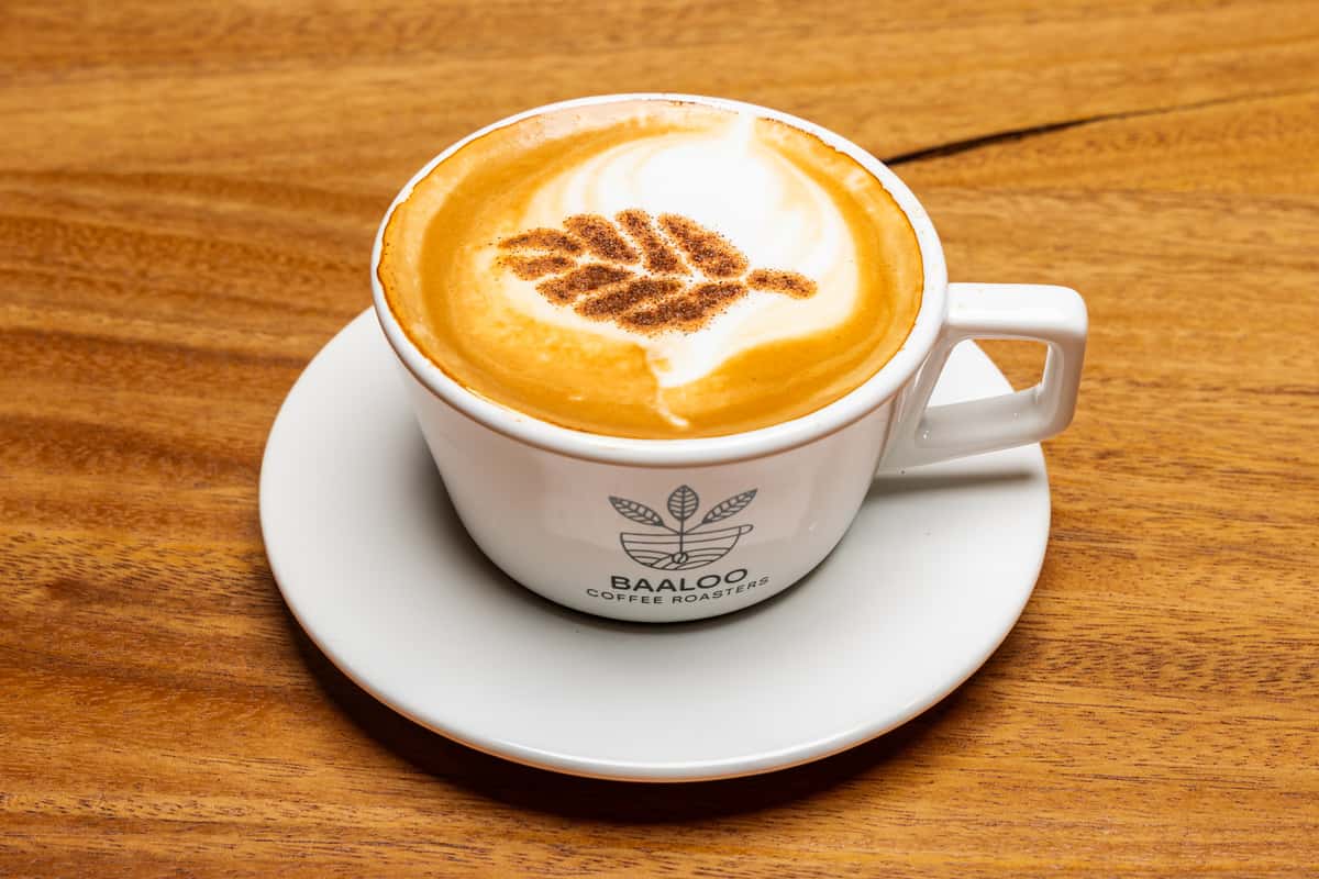 Freshly prepared cappuccino topped with creamy milk foam, cinnamon, and latte art, served in a white ceramic cup on a wooden café table.