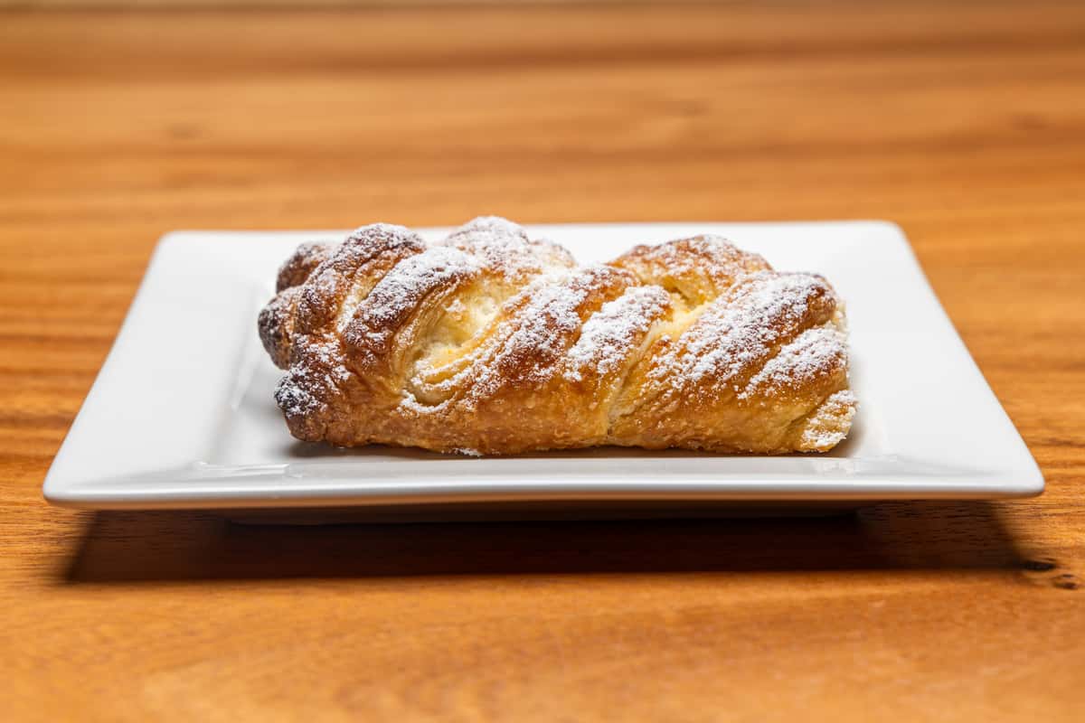 Freshly baked golden pastries topped with powdered sugar and drizzled icing, served on a rectangular plate over a warm wooden café table.