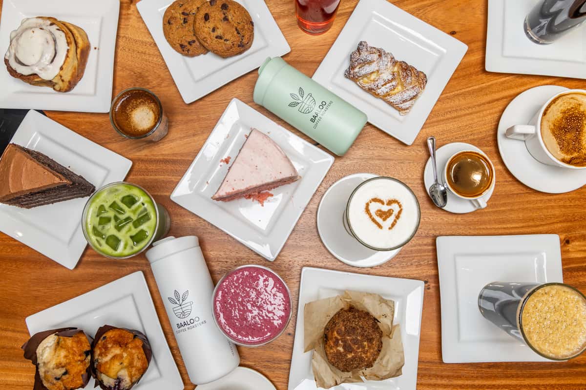 Overhead view of a delicious assortment of café breakfast and brunch dishes, including pastries, toast, eggs, fresh fruit, and specialty drinks arranged on a wooden table.