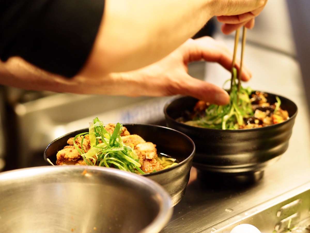 Chef garnishing two bowls of ramen with sliced green onions using chopsticks in a kitchen setting