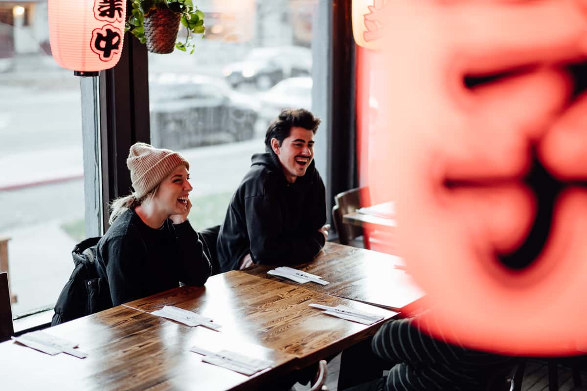 Two friends laughing at a wooden table inside a Japanese-style ramen restaurant