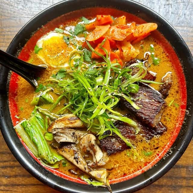 Bowl of spicy ramen with grilled beef, soft-boiled egg, kimchi, mushrooms, and green onions