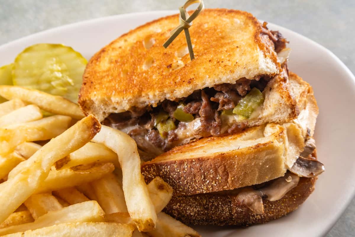 Grilled sandwich filled with beef, green peppers, and melted cheese, served with crispy fries and pickle slices on the side.