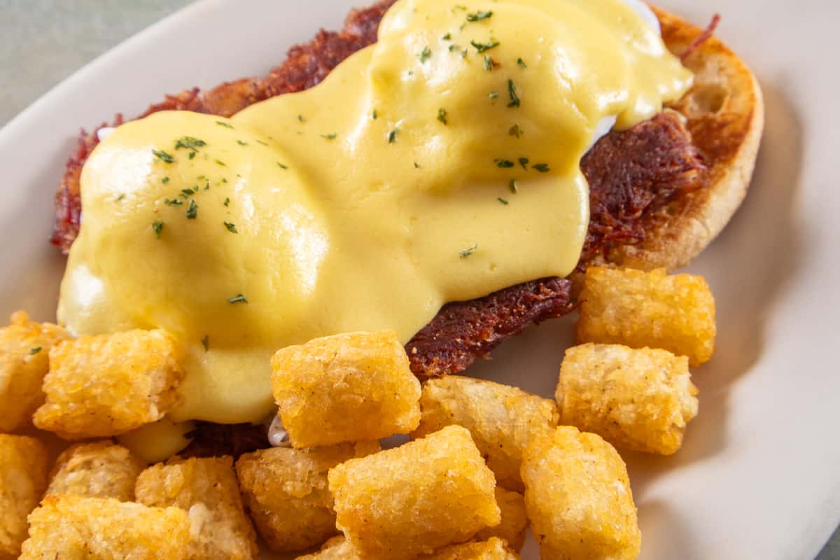 Classic eggs Benedict with hollandaise sauce, served over ham and paired with seasoned breakfast potatoes on a white plate.