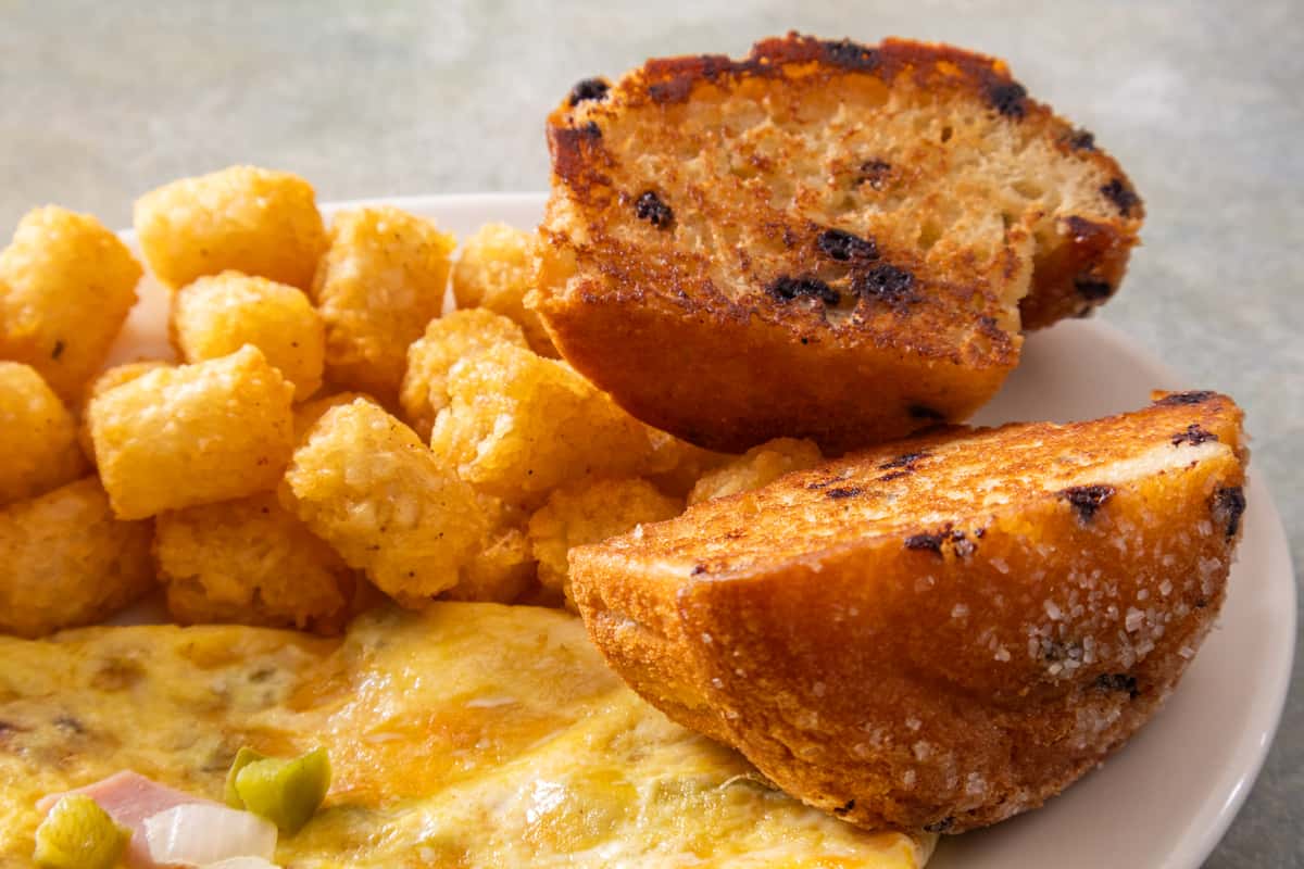 Toasted chocolate chip muffin served with tater tots and a cheesy vegetable omelet on a white plate.