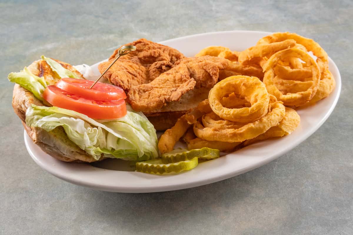Fried chicken sandwich with lettuce, tomato, and pickles served with a side of crispy golden onion rings.