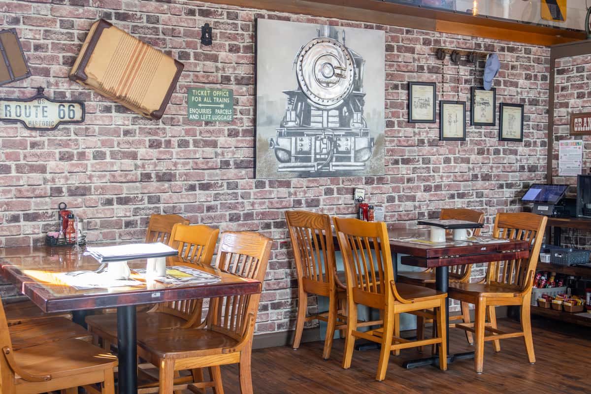 Cozy diner seating area with wooden chairs, brick walls, train-themed decor, and Route 66 and vintage signs.