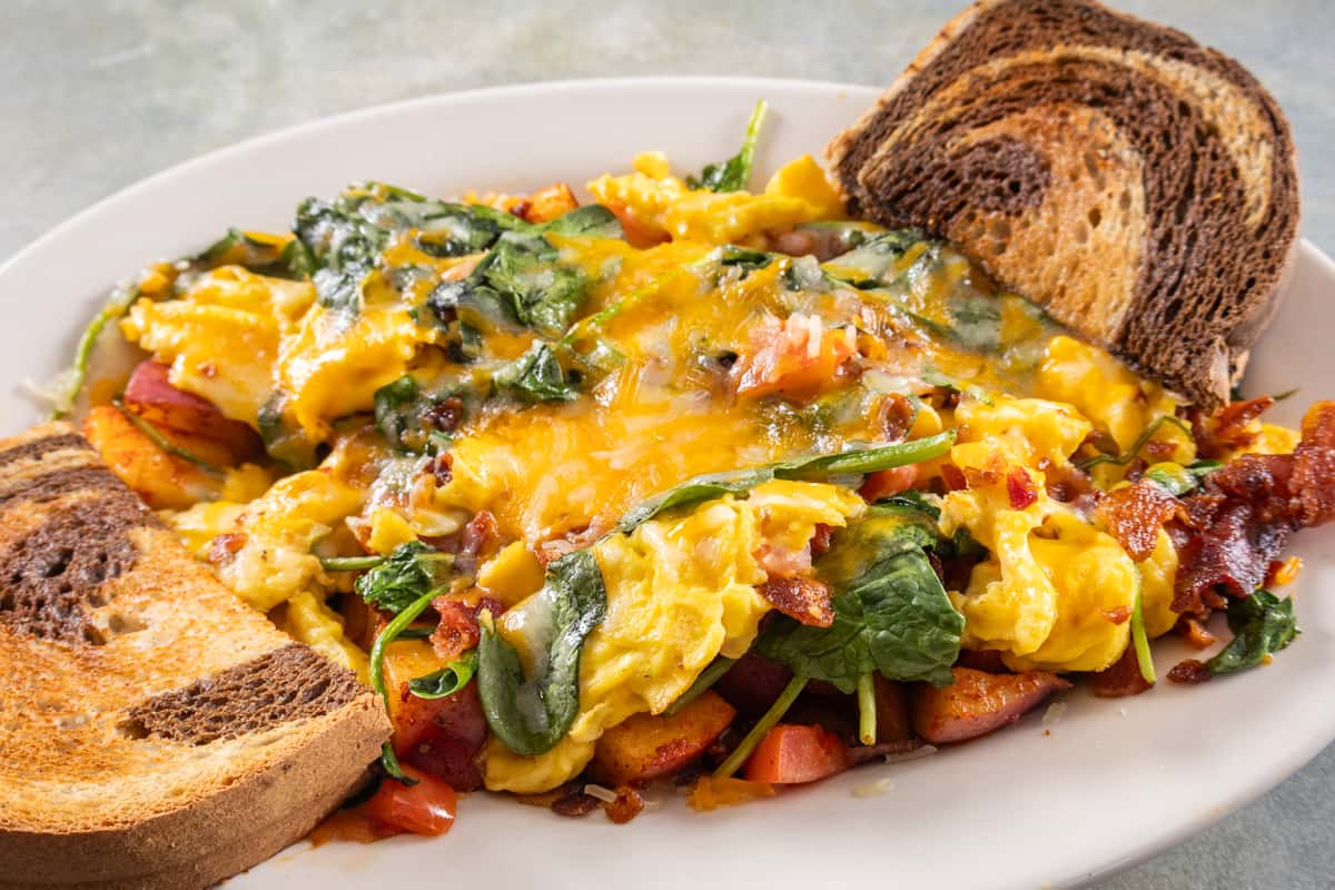 Cheesy scrambled eggs with spinach, bacon, and diced potatoes, served with slices of toasted marble rye bread.
