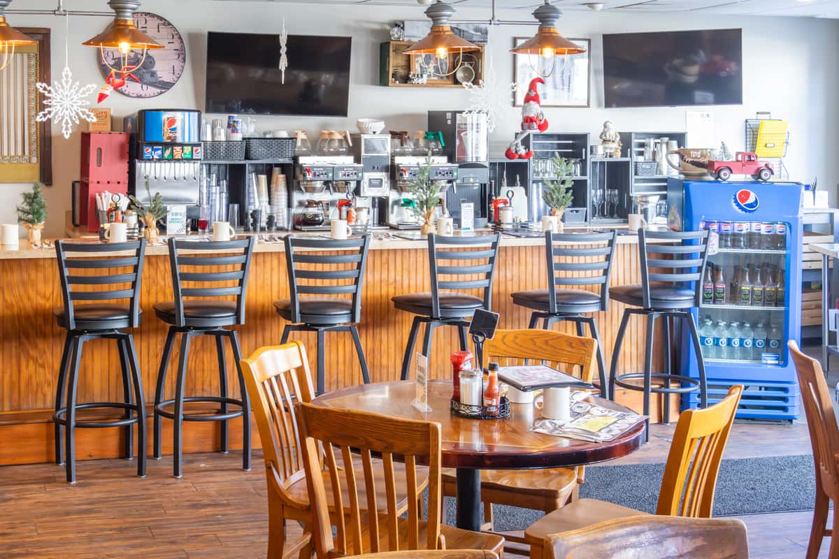 A cozy diner interior with wooden furniture, a long bar counter lined with high chairs, and festive winter decorations like snowflakes and small Christmas trees. The space includes a coffee station and multiple flat-screen TVs mounted on the wall.