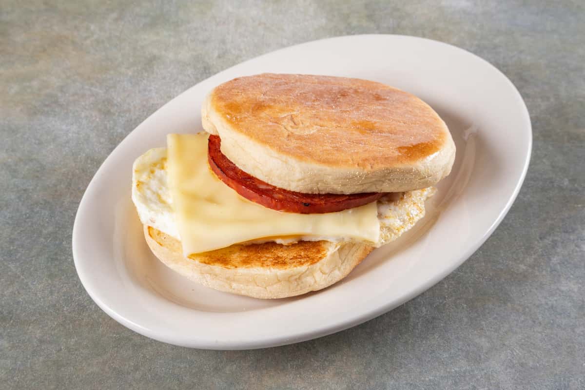 Toasted English muffin breakfast sandwich with egg, melted cheese, and a thick slice of grilled meat.