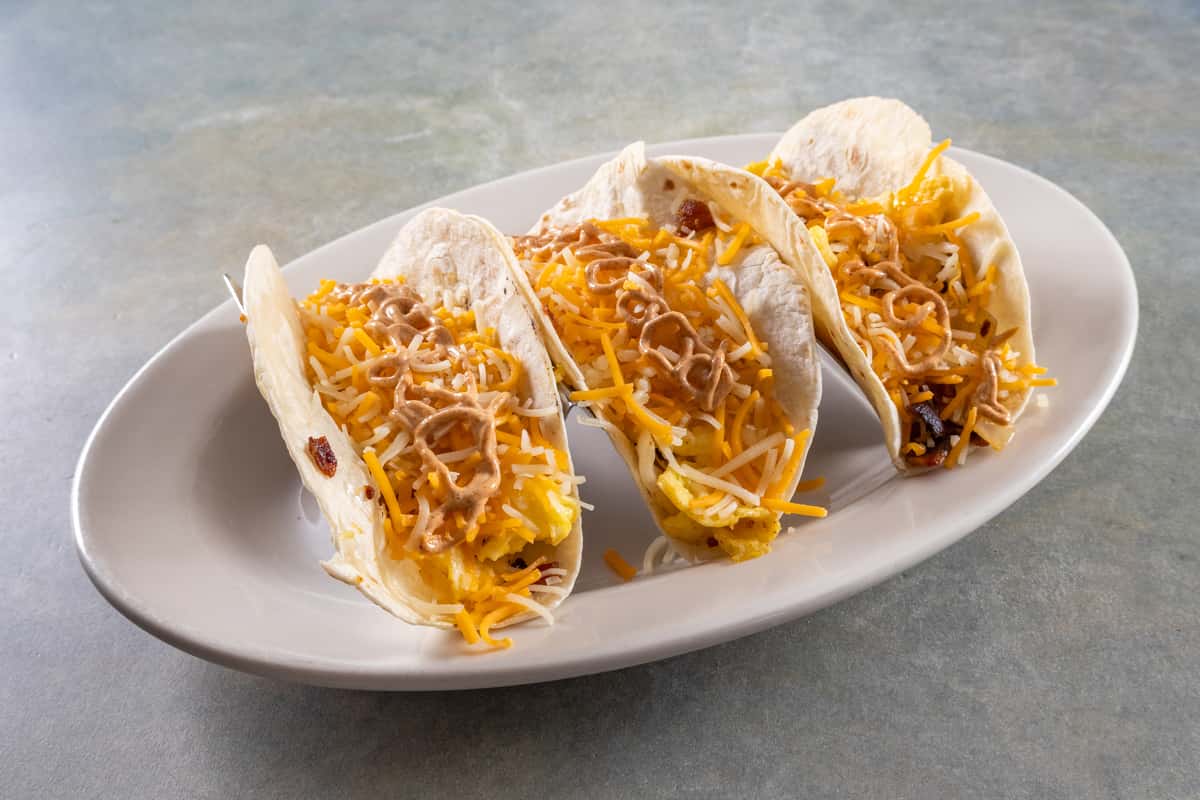 Four breakfast tacos with scrambled eggs, shredded cheese, and sauce in soft flour tortillas on a white oval plate.