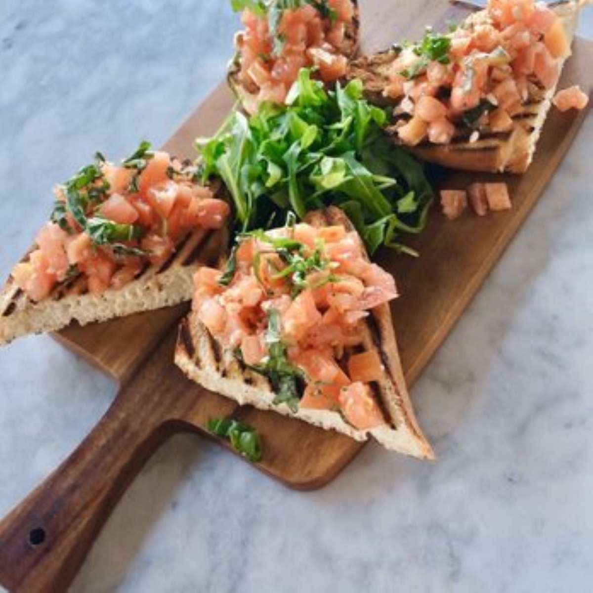 Grilled bruschetta topped with diced tomatoes and herbs, served on a wooden board with fresh arugula.