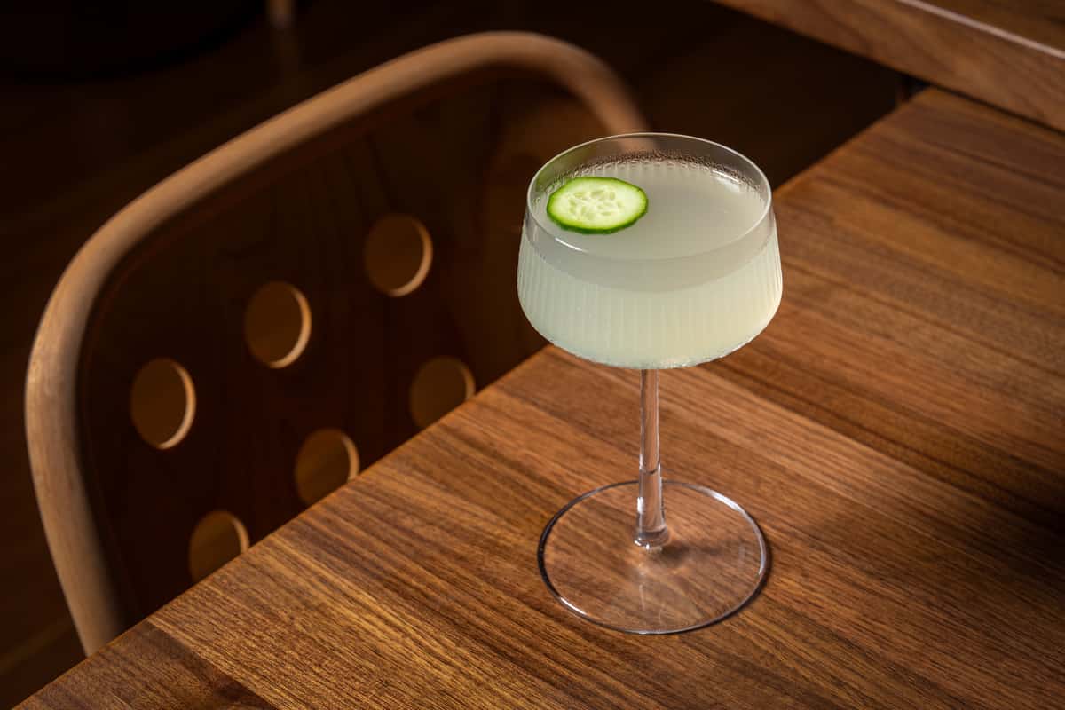 A refreshing cocktail in a crystal glass, garnished with a cucumber slice, sits on a wooden table, showcasing its pale green color and elegant presentation.
