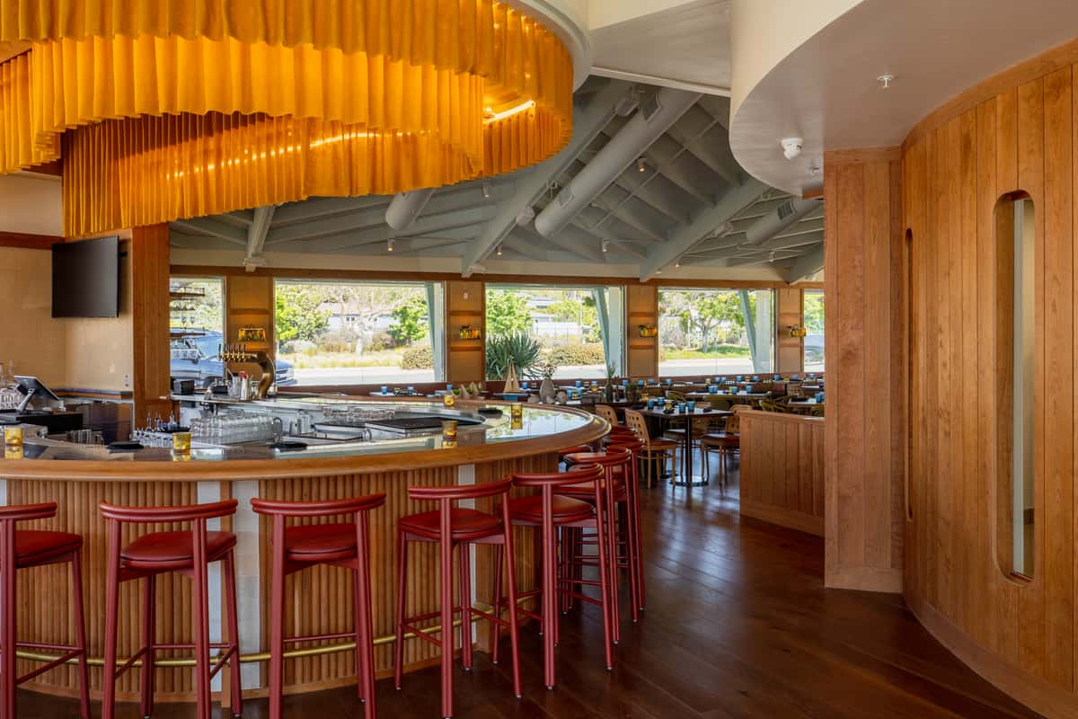 A modern restaurant interior featuring a circular bar, red stools, wooden accents, and large windows allowing natural light to illuminate the space.