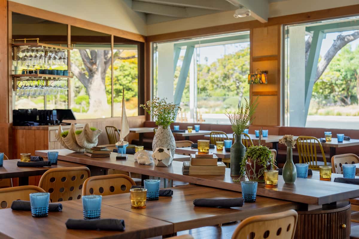 A cozy dining area featuring wooden tables set with blue and gold glasses, decorative plants, and large windows that bring in natural light and showcase the outdoor scenery.