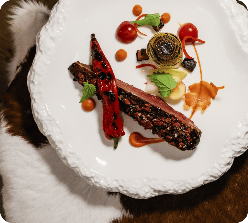 A beautifully plated dish featuring crusted meat, charred red peppers, spiraled vegetables, and vibrant sauces with mint garnishes.