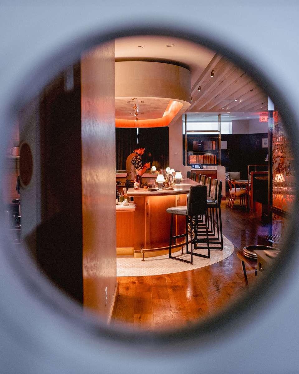 A view of a dimly lit bar from a round, slightly obscured window. A bartender is arranging drinks at the bar.