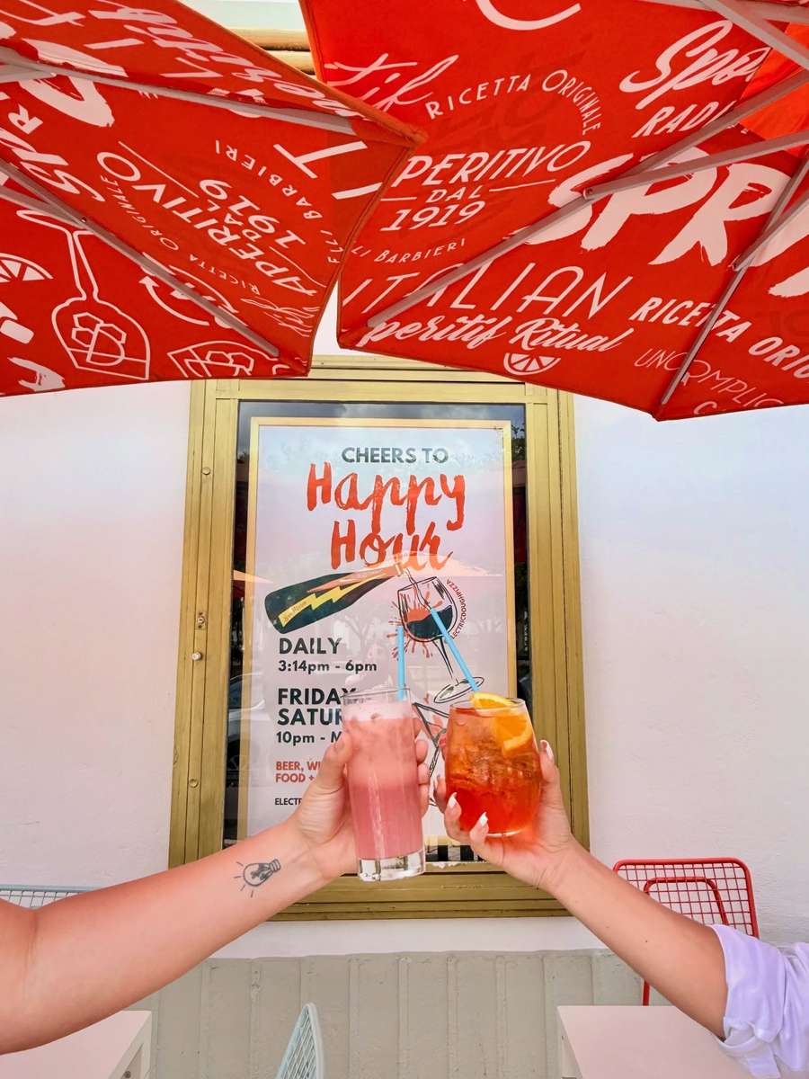 Two guests toasting drinks in front of the happy hour sign at Electric Dough Pizza Co. in San Marco Square, Jacksonville.