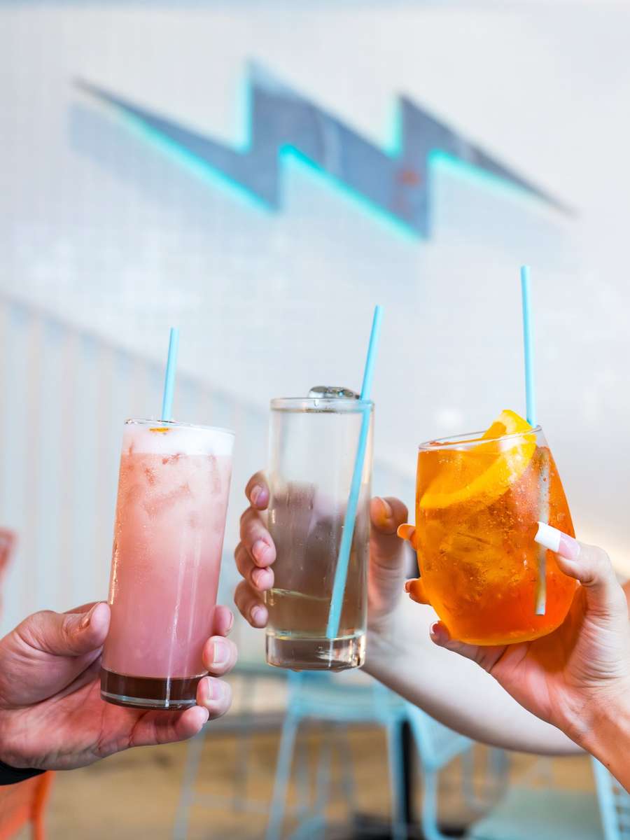 Three happy hour drinks raised in a toast in front of the neon lightning bolt wall at Electric Dough Pizza Co. in San Marco, Jacksonville.