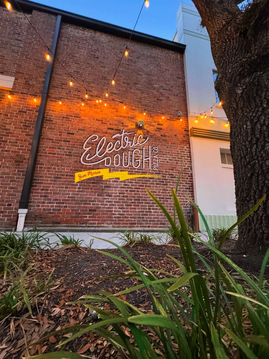 String lights over the brick side wall at Electric Dough Pizza Co. in San Marco, Jacksonville, with the painted script mark lit at dusk.