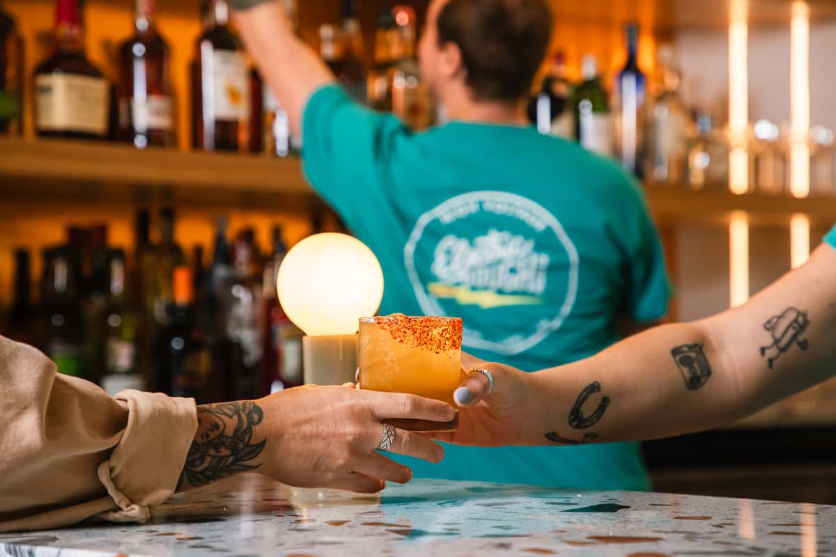 Two hands holding a cocktail at the Electric Dough Pizza Co. bar.
