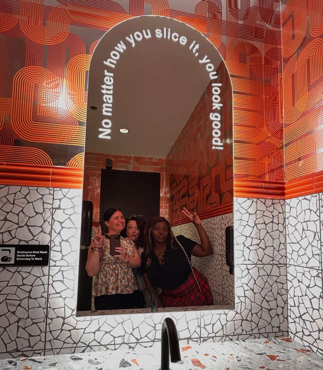 Guests posing in the Electric Dough Pizza Co. mirror that reads no matter how you slice it, you look good.