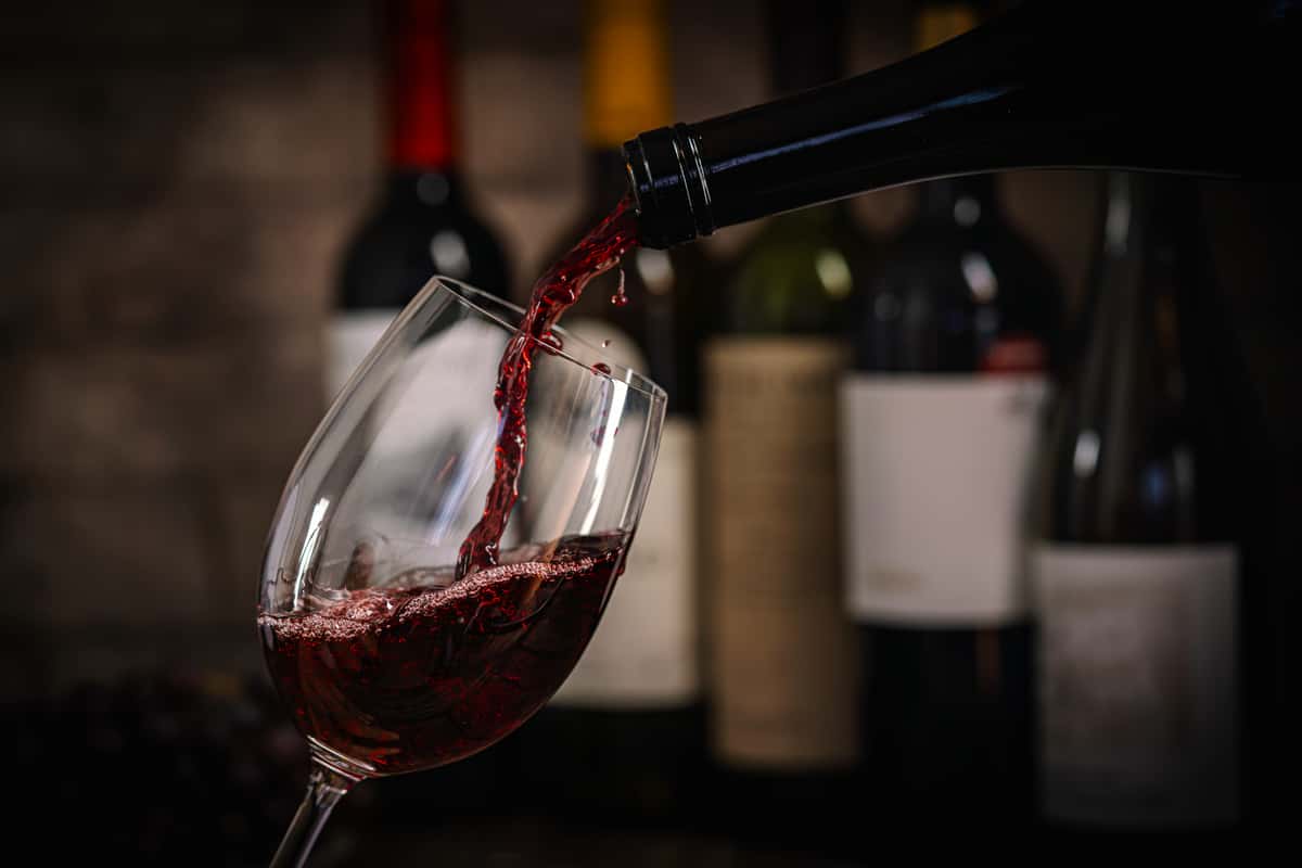 Red wine being poured into a glass in front of wine bottles.