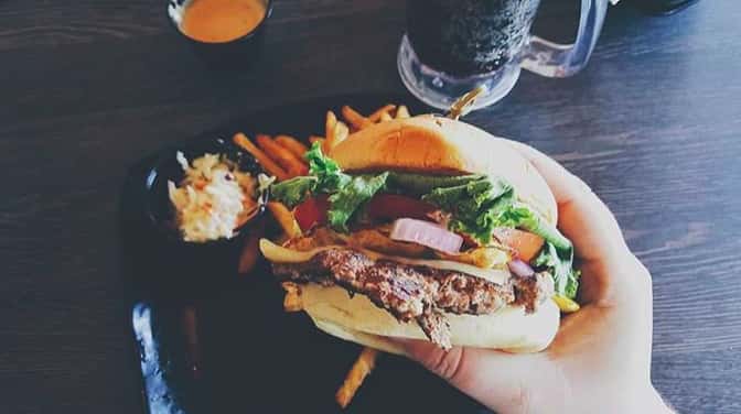 hand holding a bacon cheeseburger with tomato, lettuce, raw onion, plate of french fries and mug of root beer in background