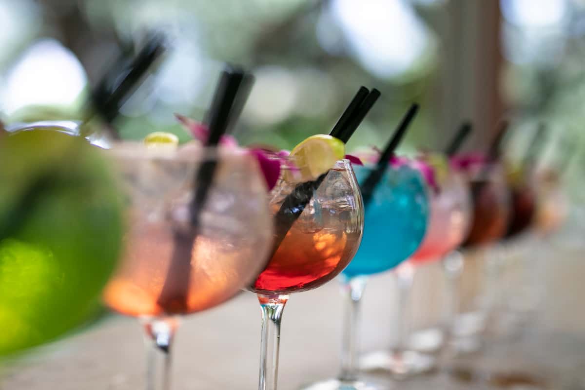 Close-up of colorful signature cocktails lined up at a restaurant bar, featuring vibrant mixed drinks with citrus garnishes, black straws, and floral accents.