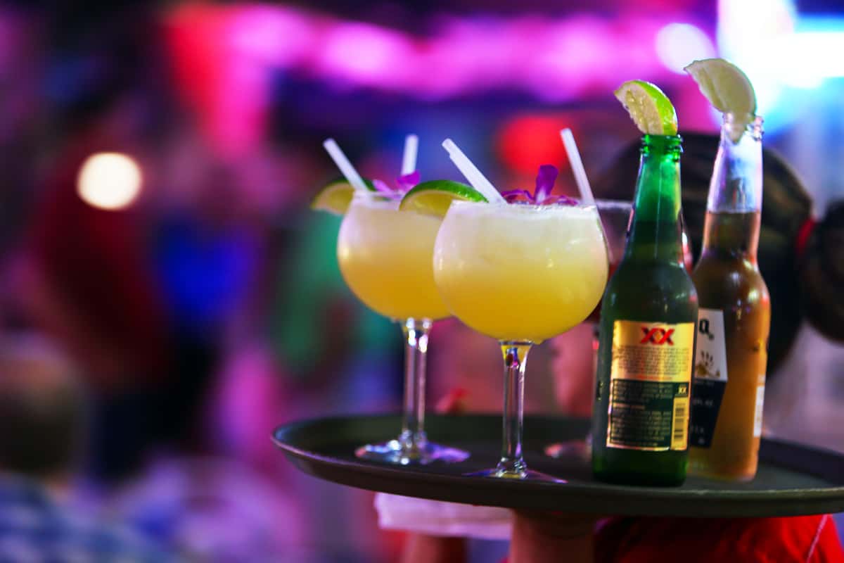 Fresh margaritas served on a tray with lime garnishes alongside Mexican beers, set against a vibrant nightclub-style bar background with colorful lighting.
