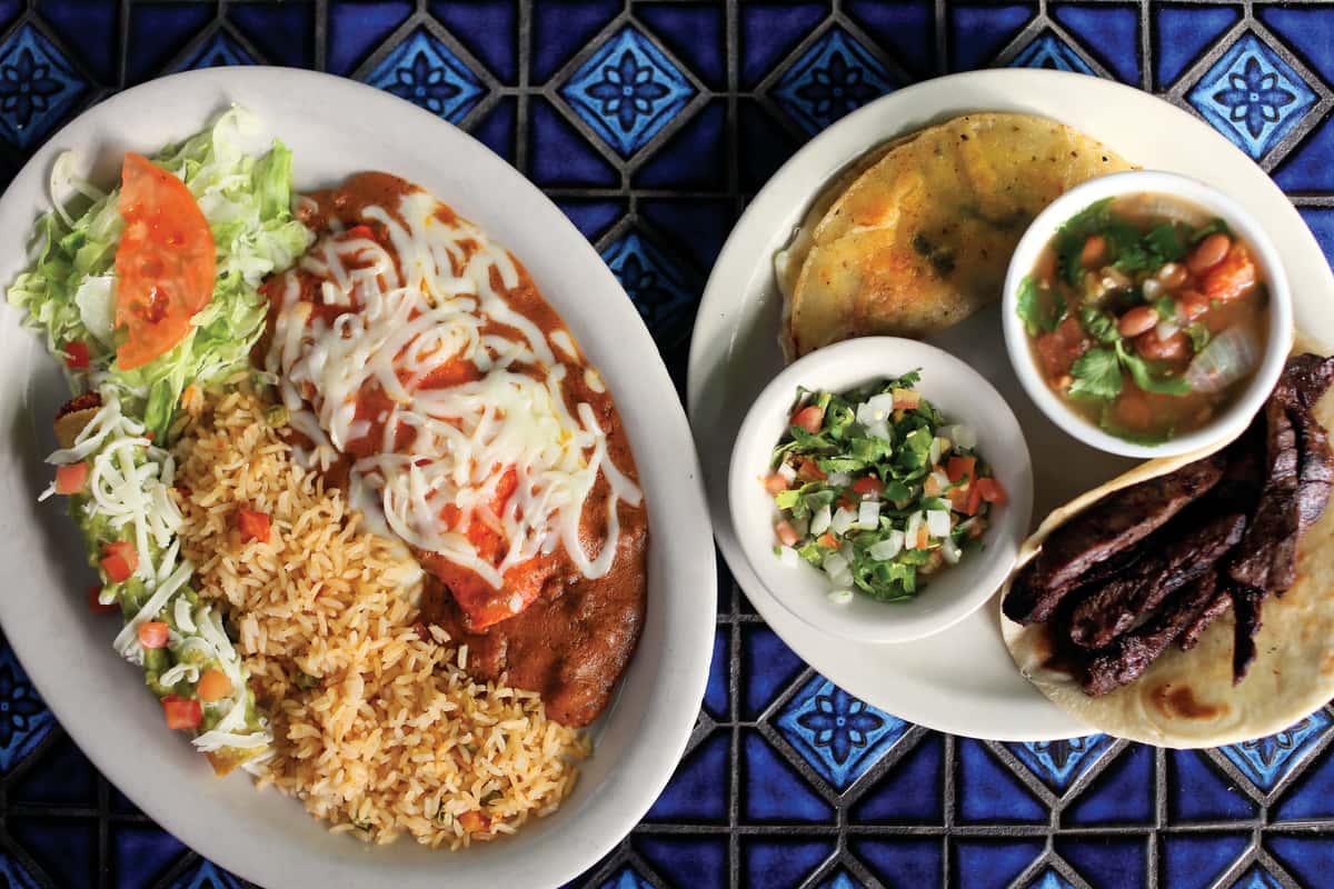 Authentic Mexican food plates featuring a cheese-topped enchilada with red sauce, Mexican rice, refried beans, fresh lettuce and tomato, plus a steak taco with pico de gallo and charro beans.