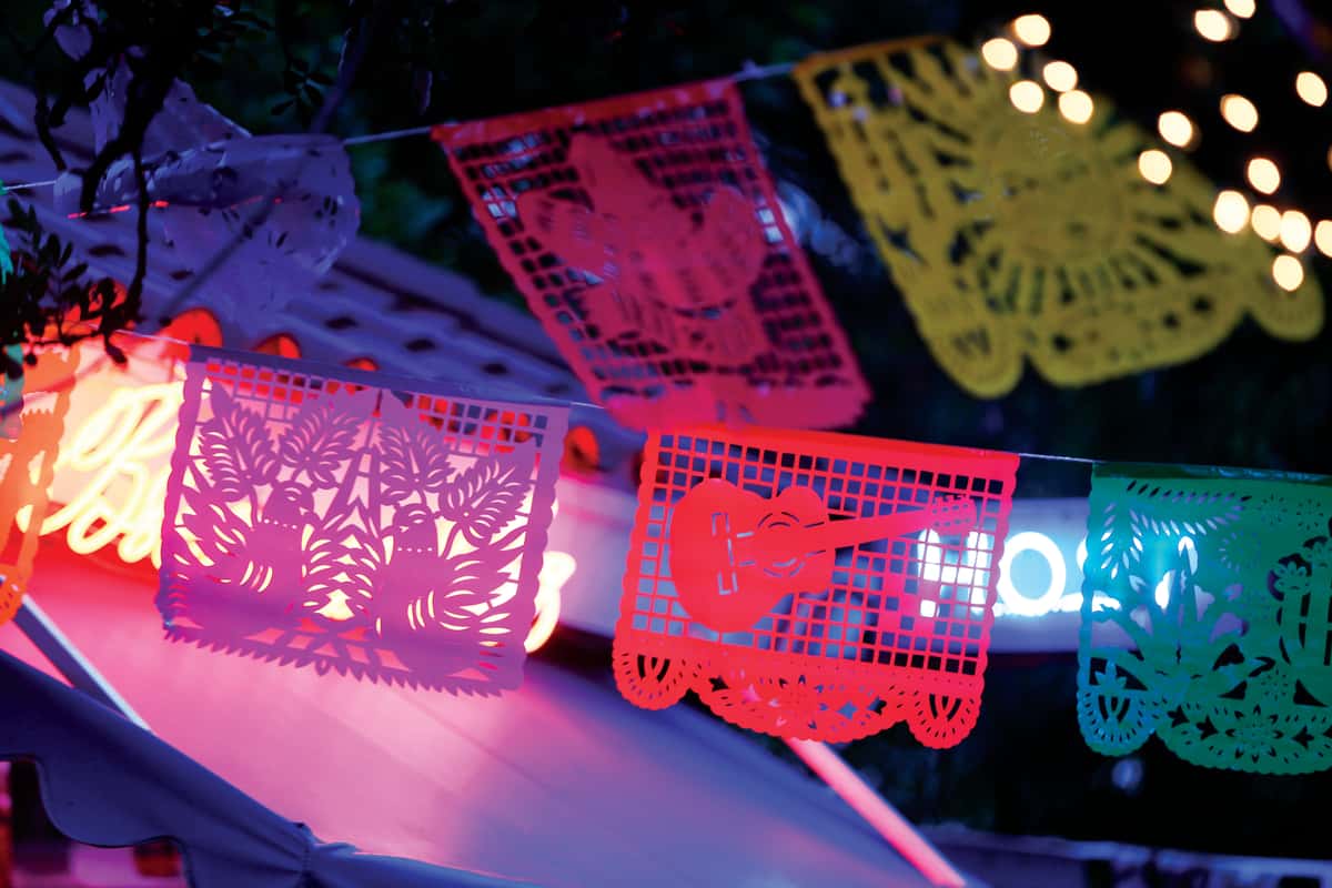 Colorful papel picado banners hanging under string lights at night, creating a festive Mexican restaurant atmosphere with glowing neon accents.