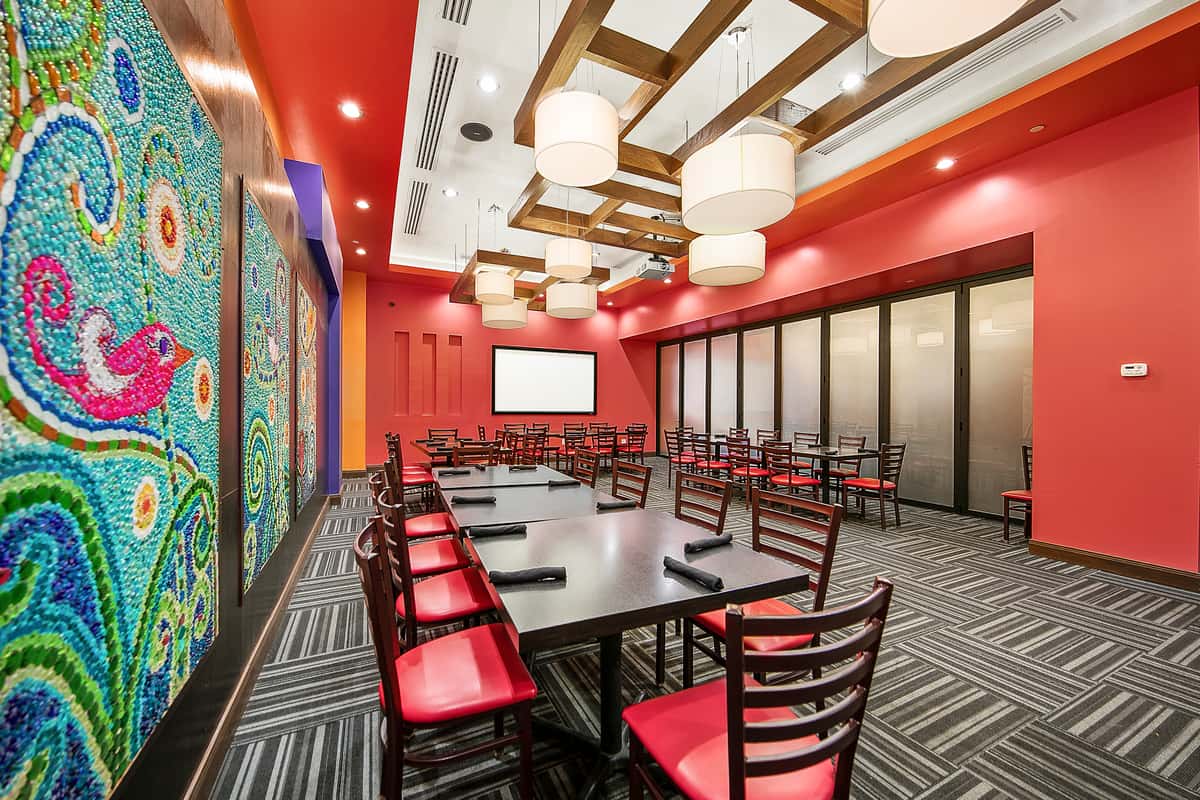 Modern private dining room in a Mexican restaurant with long tables, red accent walls, colorful mosaic art panels, wood ceiling beams, and contemporary pendant lighting.