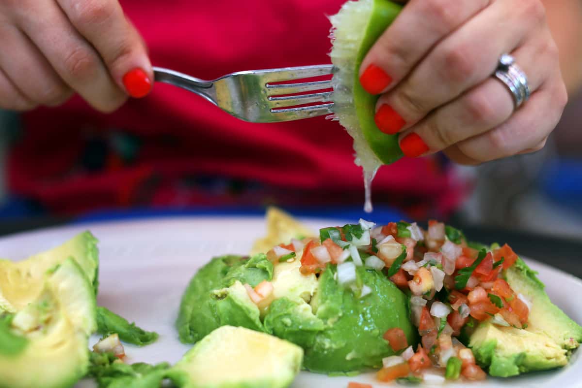 Fresh guacamole topped with pico de gallo as lime is squeezed over sliced avocado at a Mexican restaurant table.