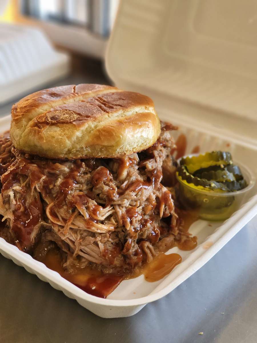 Pulled pork sandwich smothered in barbecue sauce with a soft bun, served in a takeout container with a side of pickles.