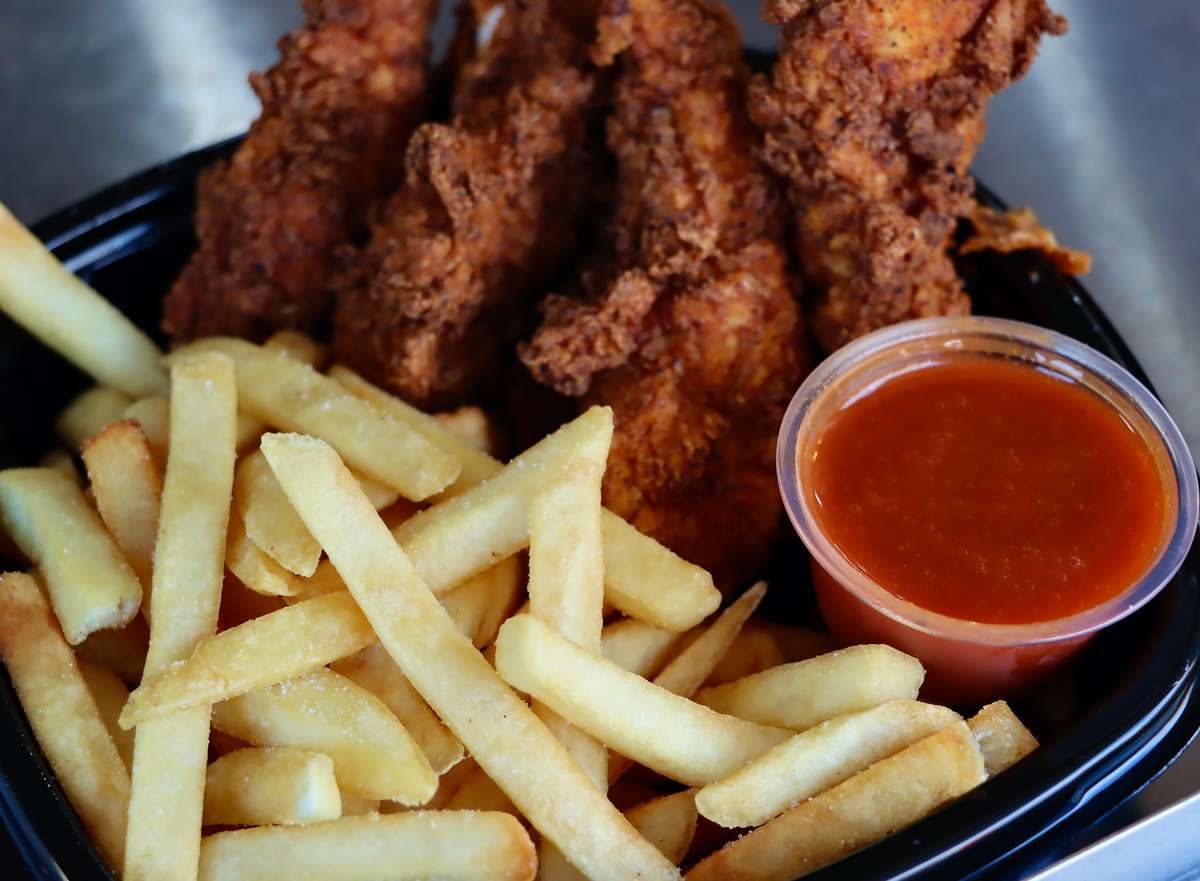 Crispy fried chicken tenders with golden French fries and a side of dipping sauce in a black takeout container.