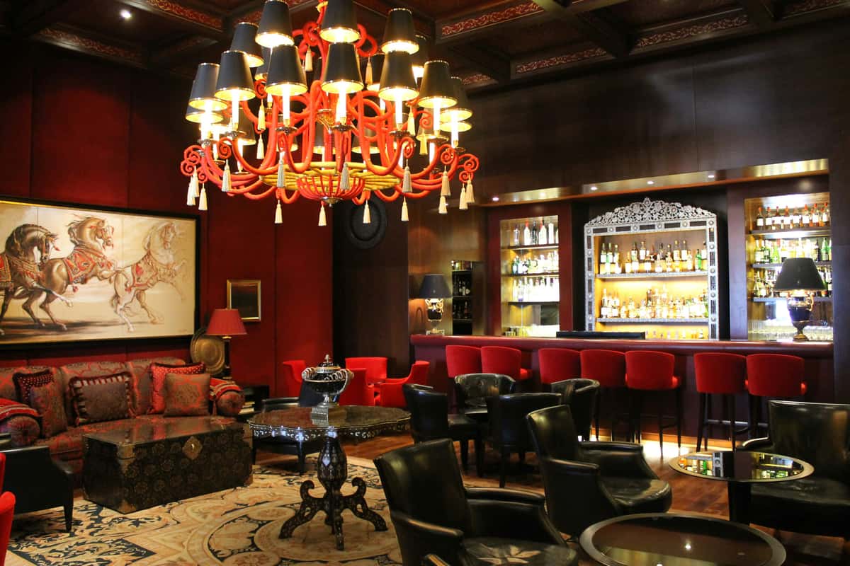 Opulent cocktail lounge with red velvet seating, a dramatic chandelier, ornate décor, and a fully stocked mirrored bar.