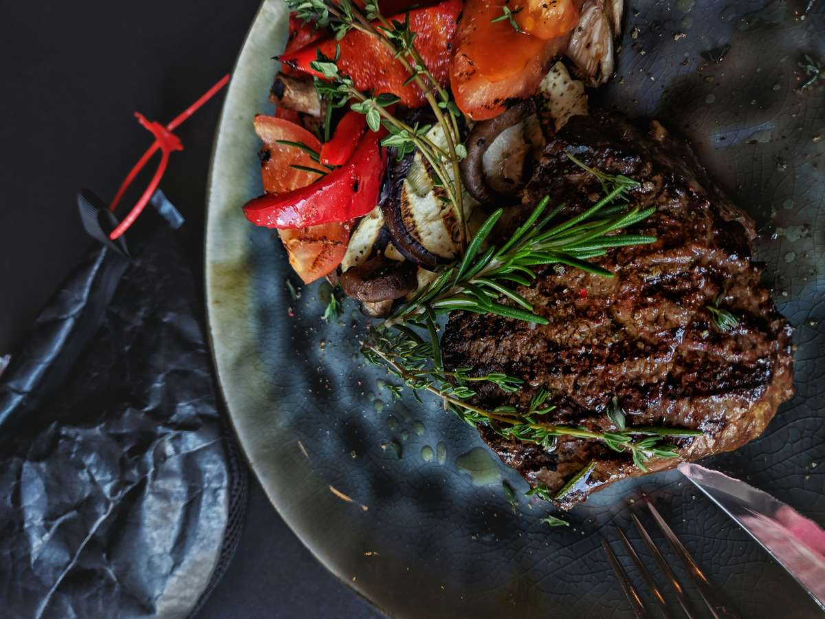 Grilled steak topped with rosemary, served with roasted red peppers, zucchini, and mushrooms on a dark rustic plate.