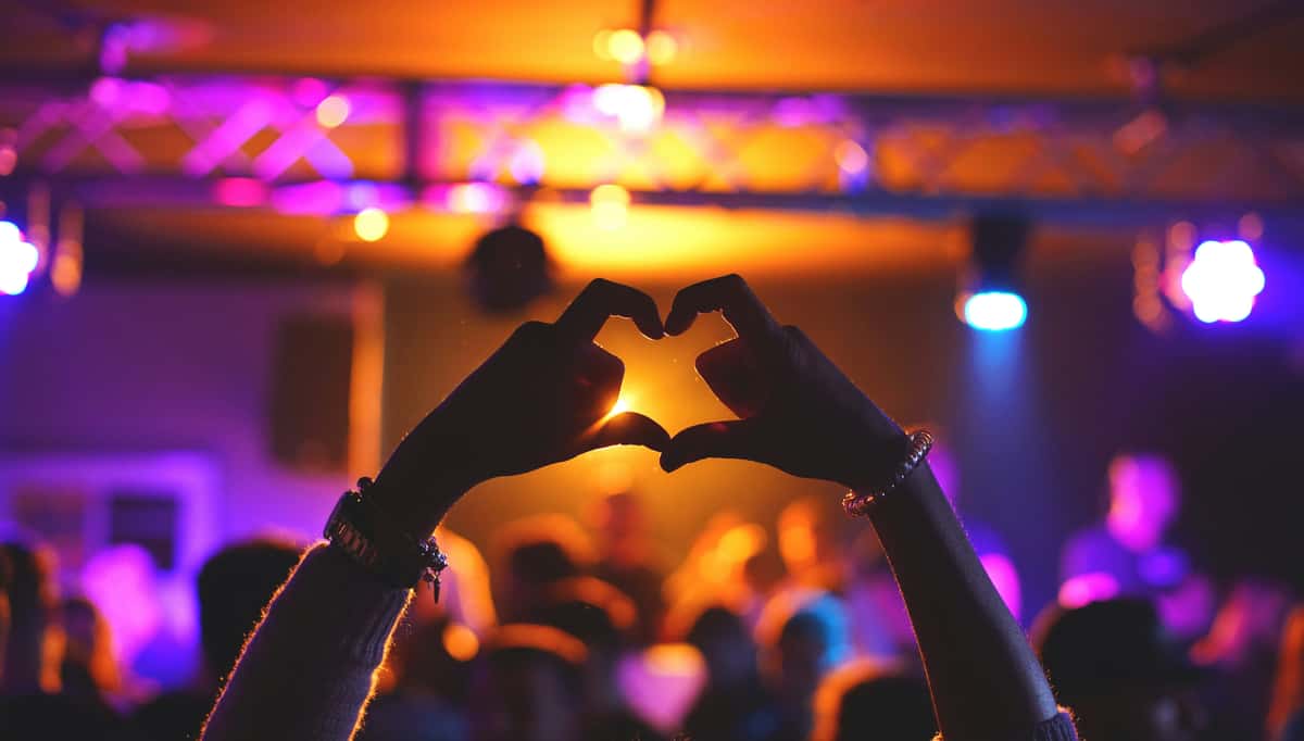 Hands forming a heart shape at a concert with colorful stage lights and a blurred crowd in the background.