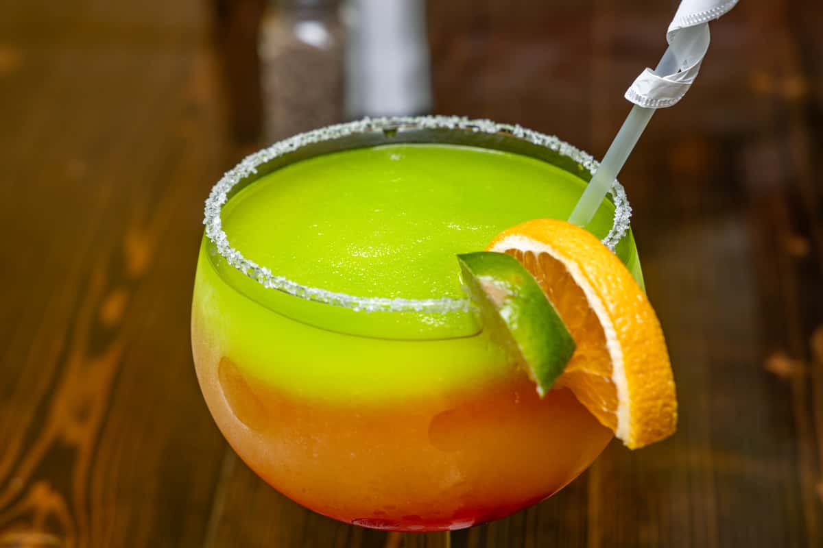 Layered frozen cocktail with vibrant green, orange, and red hues in a salt-rimmed glass, garnished with lime and orange slices.