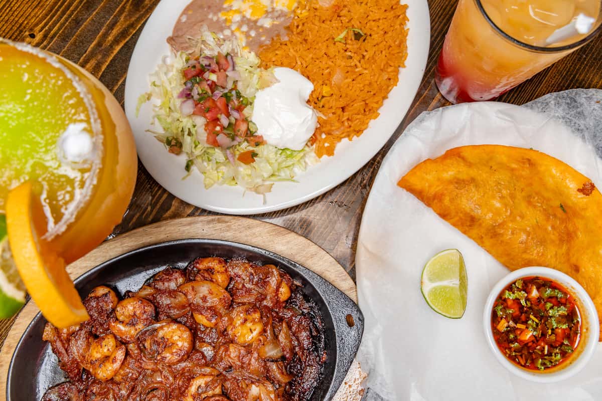 Mexican plate with shrimp and onions, rice, beans, lettuce, empanada, pico de gallo, and cocktails on a rustic wooden table.